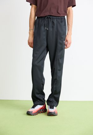 Persoană îmbrăcată în pantaloni de trening negri cu șnur și adidași albi, roșii și negri, stând pe un podea verde, împotriva unui perete alb.