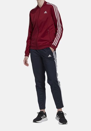 Osoba ubrana w bordową bluzę sportową Adidas zapinaną na zamek oraz granatowe spodnie sportowe Adidas z białymi paskami i czarne buty sportowe Adidas.