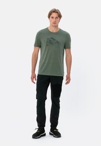 Groen sportief T-shirt met een bergafbeelding in zwart en blauw, gecombineerd met zwarte technische broek. Model staat tegen een witte achtergrond.
