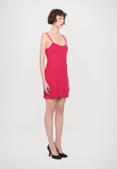 Abito rosa aderente con motivo a zig zag, spalline sottili e texture liscia, abbinato a tacchi a spillo neri.