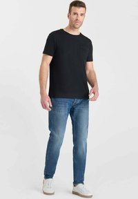 T-shirt nera a maniche corte con taschino, abbinata a jeans azzurri. Sneakers bianche con suola beige. Look casual e aderente.