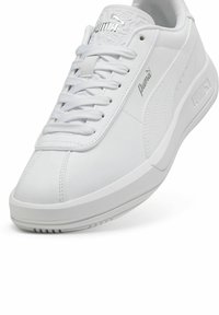 Adidași albi cu un superior sintetic neted, vârf rotund și talpă din cauciuc texturat. Caracteristicile includ șireturi și un logo ridicat pe lateral.