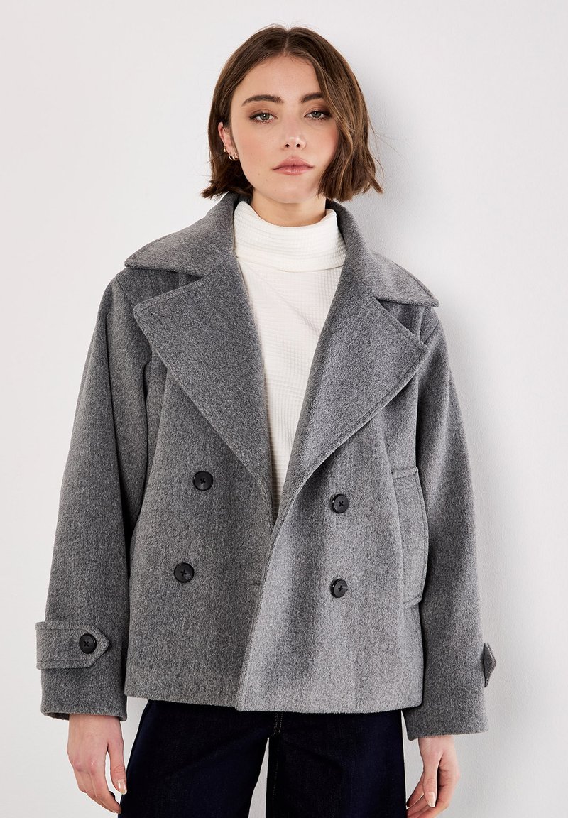 Cappotto di lana grigio doppiopetto con colletto a revers, bottoni neri, vestibilità oversize e maniche larghe, indossato sopra un dolcevita bianco.