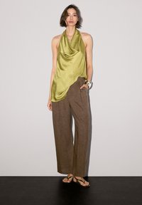 Top en satin vert olive avec un design drapé, associé à un pantalon en lin marron ample. Le mannequin porte des tongs noires et un bracelet en argent.