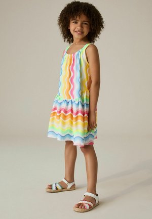 Mladé dievča s kučeravými vlasmi, oblečené v bezrukávových šatách s dúhovými vlnitými pruhmi a bielymi sandálmi, stojí na jednoduchom pozadí.