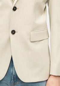 Μπεζ σακάκι με μαύρα κουμπιά και τσεπάκι με καπάκι, φορεμένο πάνω από μπλε τζιν, εμφανίζοντας το κάτω μέρος του κορμού και το δεξί χέρι.