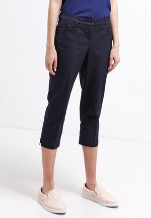 Person wearing navy cropped pants with front pockets, slit hems, a navy top, and light pink slip-on shoes standing on a white background.