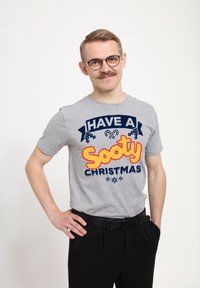 Šedé tričko s krátkým rukávem s barevným grafickým potiskem s nápisem "Přejeme kouřové Vánoce." V kombinaci s černými kalhotami a páskem.