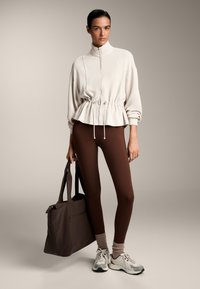 Lys beige zip-up sweatshirt med indsnævret talje, brune leggings, beige sneakers og en brun tote bag. Blødt stof, afslappet pasform og ribbede detaljer.