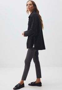 Camicia nera oversize, jeans slim-fit grigi, mocassini neri con nappe. Tessuto morbido, design minimalista, dettaglio con bottoni laterali sulla camicia.