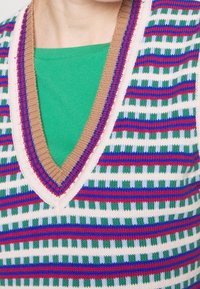 Gilet en maille avec décolleté en V. Base blanche avec des rayures horizontales rouges, bleues, vertes et violettes, avec une bordure côtelée marron.