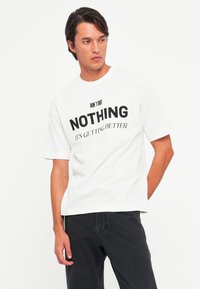 Mann som har på seg en hvit T-skjorte med svart tekst «AIN'T GOT NOTHING IT’S GETTING BETTER» og svarte bukser, står med den ene hånden i lommen.
