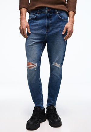 Man wearing blue ripped skinny jeans, brown long-sleeve shirt, and black chunky sneakers, standing on white background.