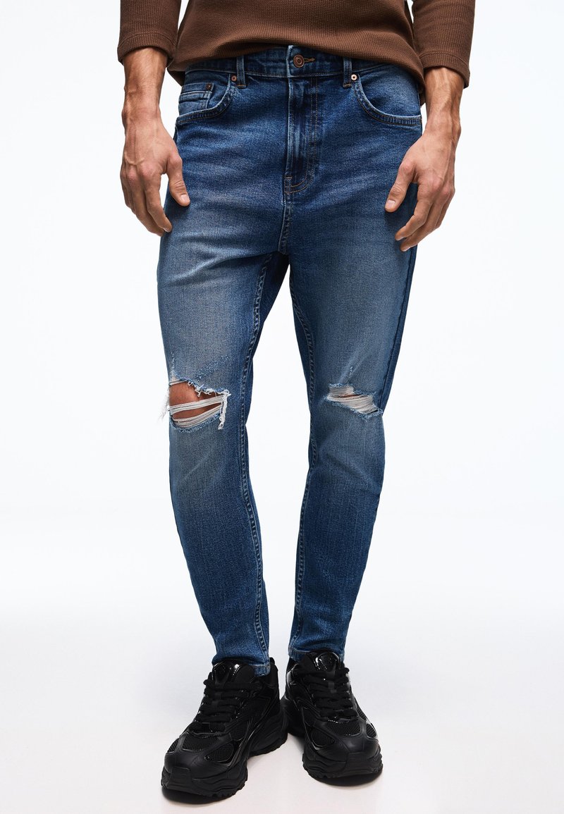 Bărbat purtând blugi skinny albaștri cu rupturi, cămașă maro cu mâneci lungi și adidași negri masivi, stând pe fundal alb.