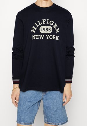 Osoba ubrana w granatową koszulkę z długim rękawem z napisem "HILFIGER 1985 NEW YORK" oraz jasnoniebieskie szorty dżinsowe na jednolitym tle.