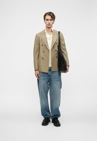 Νεαρός άνδρας στέκεται φορώντας καφέ διπλόκληρο σακάκι, κρέμ πουκάμισο, χαλαρές μπλε τζιν παντελόνες, μαύρα παπούτσια και κρατώντας μια μαύρη τσάντα ώμου.