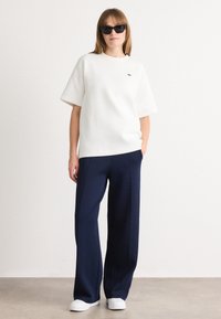 White short-sleeve top with a green logo, paired with navy wide-leg trousers. Model wears black sunglasses and white sneakers. Flat background.