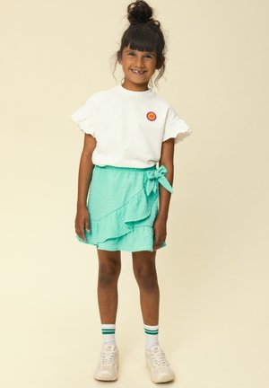 Glimlachend meisje met een hoge knot, gekleed in een wit shirt met gerimpelde mouwen en een turquoise wikkelrok, witte sneakers en witte sokken met groene strepen.