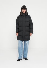 Zwarte pufferjack met een hoge hals, met brede horizontale secties, gecombineerd met lichtblauwe jeans en zwarte instapschoenen. Gladde textuur.
