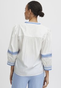 Blouse blanche avec des accents brodés bleus sur les manches et le col. Présente des manches bouffantes et un dos froncé pour un ajustement décontracté.
