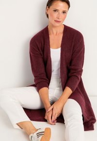 Burgundyröd lång kofta med öppen framsida och trekvartsärmar, bärs över en vit topp och kombineras med vita byxor och sportiga skor.