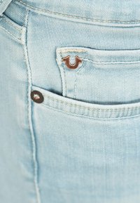 Jeans en denim bleu clair avec un petit logo couleur cuivre sur la poche arrière, avec des coutures visibles et un design standard de poche arrière.