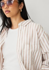 Beige og hvidstribet linnedskjorte med knaplukning foran, krave og venstre brystlomme, båret over en hvid t-shirt. Oppustede solbriller og gyldne øreringe.