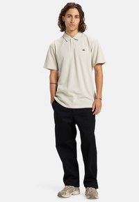 Lys beige polo skjorte med krave, prydet med et lille sort logo, sat sammen med sorte bukser og mønstrede sneakers. Casual pasform og tekstur.