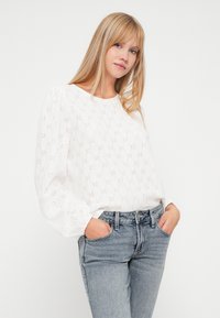 Biela blúzka s kvetinovým výrezovým vzorom, okrúhlym výstrihom a nadýchanými dlhými rukávmi. V kombinácii so svetlomodrými džínsy s vysokým pásom.