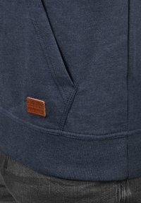 Felpa blu navy con tessuto a trama, orlo a costine e un'etichetta in pelle marrone cucita sul lato inferiore, dotata di una tasca laterale funzionale.