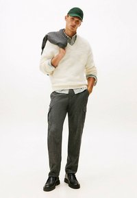 Sweter kremowy z domieszką wełny, zielona czapka, pasiasta koszula na guziki, szare spodnie typu cargo oraz czarne skórzane botki. Casualowa, warstwowa stylizacja z teksturą.