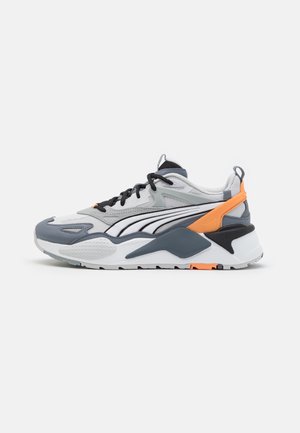 Chaussure de sport grise, blanche et orange avec des lacets noirs et une semelle épaisse, vue de profil sur un fond clair.