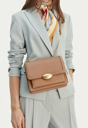 Žena ve světle šedém obleku, která nese béžovou koženou kabelku přes rameno se zlatou sponou a má kolem krku vzorovaný hedvábný šátek.