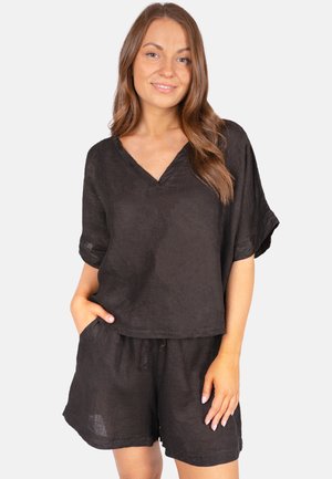 Jeune femme aux longs cheveux bruns portant un haut noir ample à col en V et un short assorti, debout avec une main dans la poche.