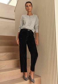 Harmaa pilkullinen villatakki nappeilla yhdistettynä korkeavyötäröisiin mustiin ruutuhousuihin ja harmaisiin kuviollisiin slingback-korkokenkiin. Taustalla puinen portaikko.