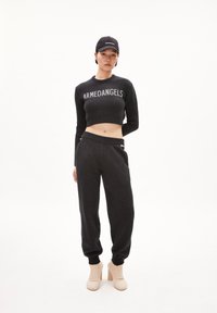 Suéter negro recortado con el logo "ARMEDANGELS", combinado con pantalones jogger negros. La modelo lleva botines de punto beige hasta el tobillo. Diseño simple y minimalista.