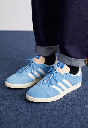 Osoba nosząca niebieskie zamszowe buty Adidas Gazelle z białymi paskami, beżowym językiem, czarnymi skarpetkami i podwiniętymi ciemnymi dżinsami na niebieskim dywanie.