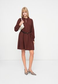 Καφέ φόρεμα με μακριά μανίκια και λαιμόκοψη τύπου κλειδώματος, που είναι μαζεμένο στη μέση με μια μαύρη ζώνη. Ζευγάρι ανοιχτόχρωμες γκρι γόβες slingback και μια μικρή λευκή τσάντα.