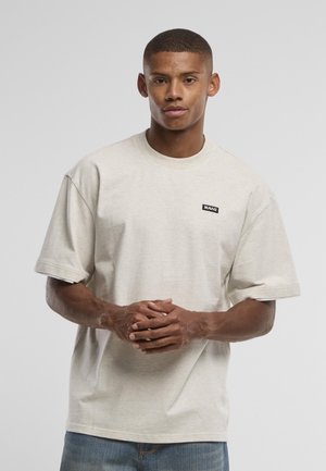 Fiatal férfi, rövid hajjal, bézs, túlnagy pólót visel, amelyen kis fekete logó van a mellkasán, összekulcsolt kézzel áll.