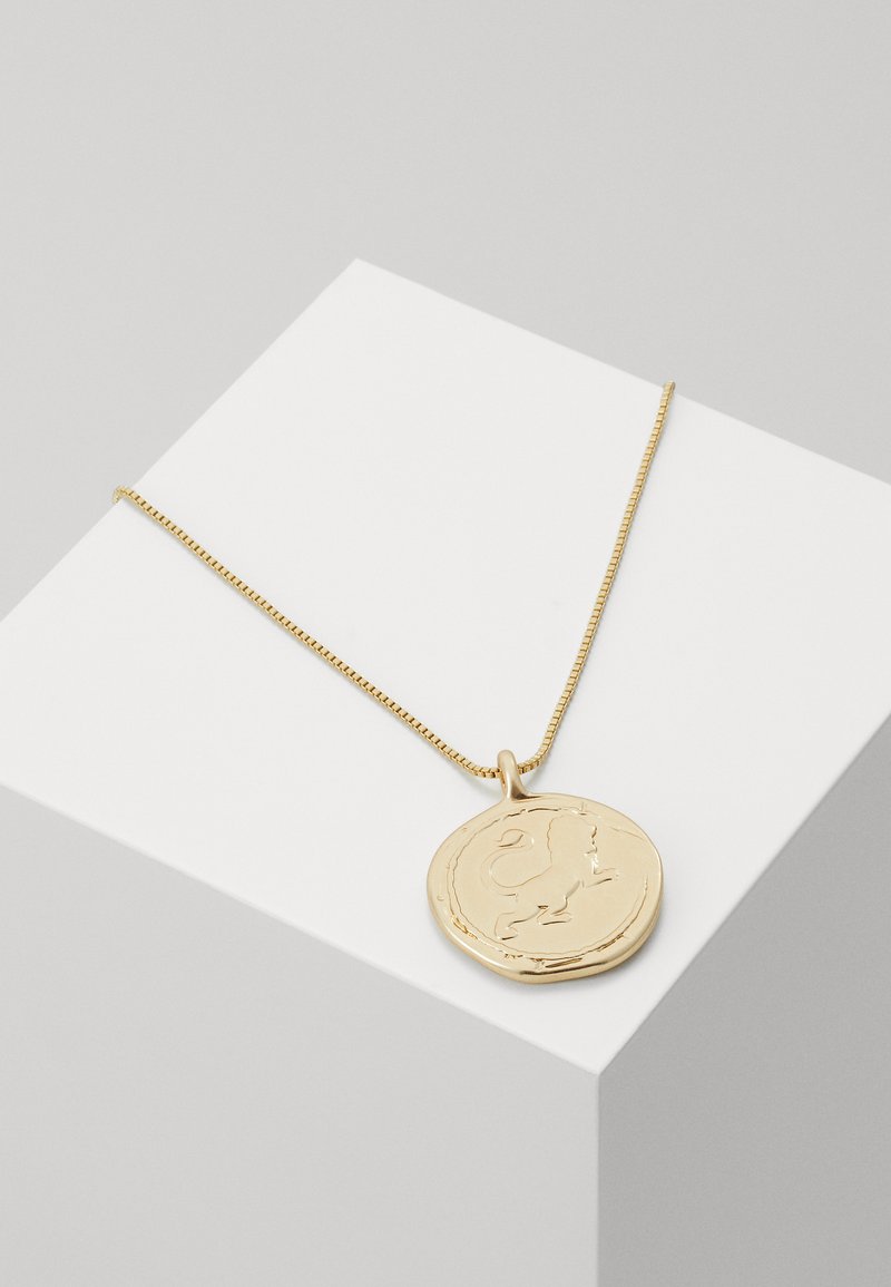 Collar con colgante en tono dorado que presenta un charm redondo con un diseño de león. La cadena es de estilo delicado y retorcido. Superficie lisa y reflectante.