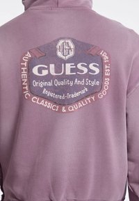 Felpa a maniche lunghe color lavanda con una grande grafica stampata sulla schiena, caratterizzata da un logo, testo e un look vintage consumato.