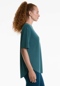 Haut teal à manches courtes en tissu lisse et extensible avec un ourlet arrondi, associé à des leggings navy, vu de côté.