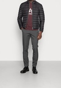 Černá puffer bunda s horizontálním prošíváním, vínové tričko s bílou grafikou, šedé přizpůsobené kalhoty a černé boty. Jednoduchý, moderní design.