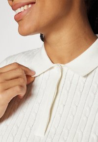 White knitted polo shirt with a textured pattern, featuring a flat collar and button closure. Hand holding the collar at the neck area.