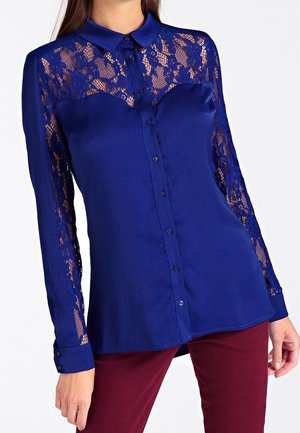 Femme portant un chemisier bleu boutonné avec des panneaux en dentelle sur les épaules et les manches, associé à un pantalon rouge foncé, debout devant un fond blanc.