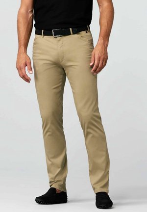 Mann trägt schlanke beige Hose mit schwarzem Gürtel und schwarzen Schuhen, steht vor einfachem hellem Hintergrund.