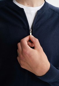 Marineblauer Reißverschluss-Pullover mit Rundhalsausschnitt und gerippten Bündchen. Ausgestattet mit einem silbernen Reißverschluss und strukturiertem Strickstoff. Wird über einem weißen Hemd getragen.