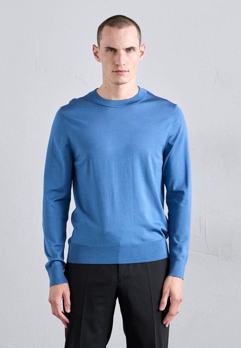 Šviesiai mėlynas megztinis su apvalia iškirpte, ilgomis rankovėmis ir giriamaisiais kraštais. Paprastas dizainas su lygiu paviršiumi, derinamas su juodomis kelnėmis.