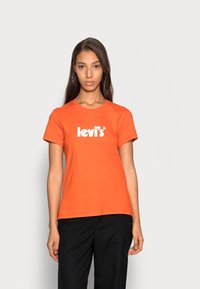 Camiseta de algodón naranja con cuello redondo y mangas cortas, que presenta el logo en blanco de "levi's" centrado en el pecho. Pantalones negros lisos.