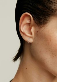 Pendiente de aro de plata con pequeñas piedras de zirconia cúbica transparentes, diseño curvado, usado en el lóbulo de la oreja. Tono de piel visible cerca del pendiente.